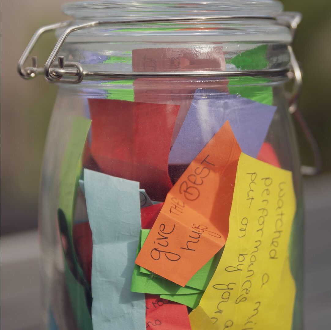May contain a glass jar filled with colorful slips of paper, some with handwritten messages like "give the best hugs".
