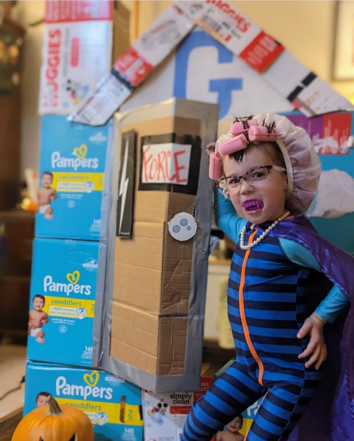 May contain a person wearing a shower cap, hair rollers, vampire teeth, and a cape, standing next to stacks of diaper boxes.