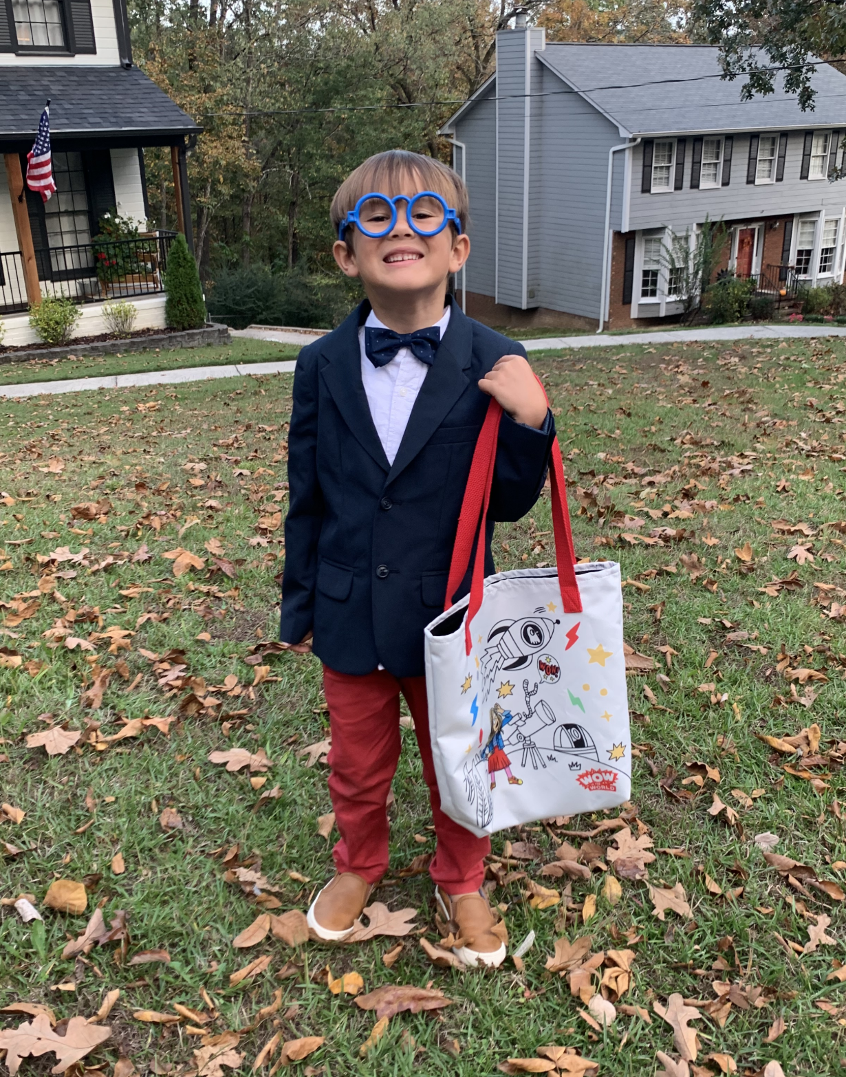May contain a person wearing blue glasses, a navy blazer, and a bow tie, holding a patterned tote bag on a lawn.